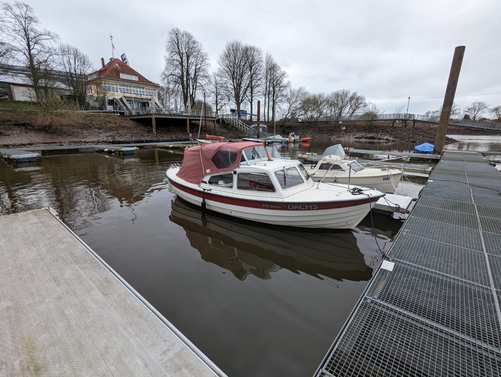 Motorboot liegt an einem Steg in einem ruhigen Hafenbecken und wird für die praktische Ausbildung zum Sportbootführerschein Binnen und See genutzt. Das Schulungsboot ist festgemacht, im Hintergrund sind weitere Stege, Gebäude und winterliche Bäume zu sehen.