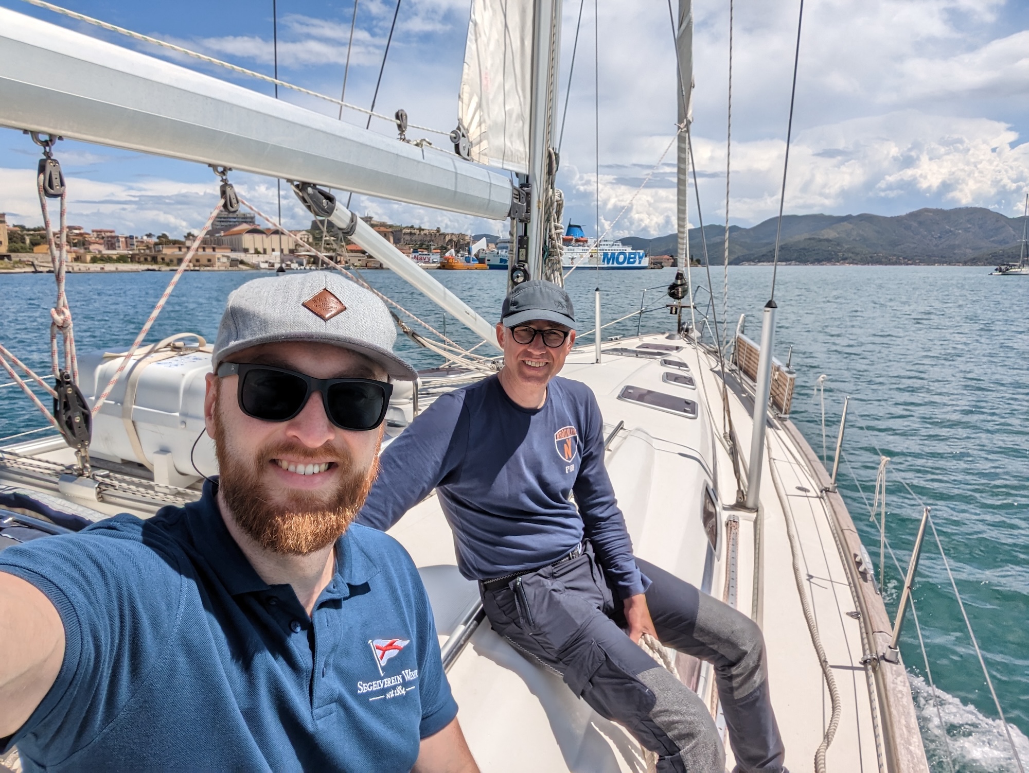 Zwei Personen sitzen an Deck einer Segelyacht während eines SSS-Praxistörns auf See. Die Yacht fährt bei ruhigem Wetter entlang einer Küste, im Hintergrund sind Hafenanlagen und Berge zu sehen. Die Szene steht für die praktische Ausbildung zum Sportseeschifferschein.