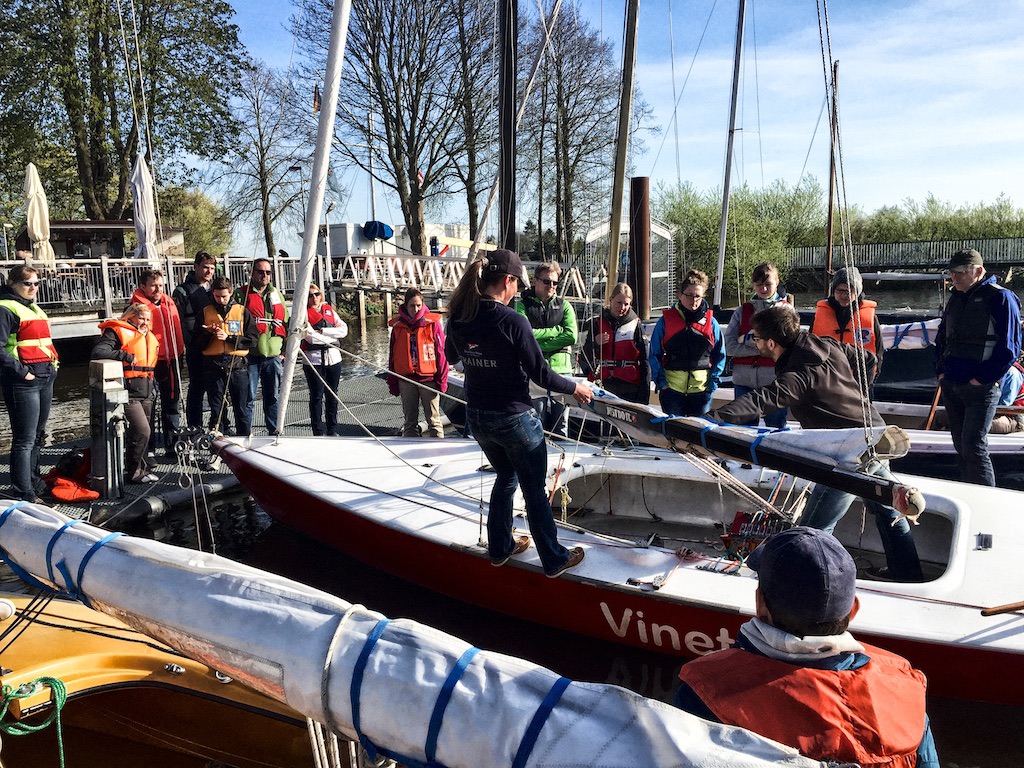 Mehrere Segelschülerinnen und Segelschüler nehmen an einer Praxisausbildung im Sommer teil und bereiten ihre Jollen am Steg vor. Unter Anleitung lernen sie grundlegende Segelmanöver und den sicheren Umgang mit Boot und Segeln beim richtigen Segeln lernen.