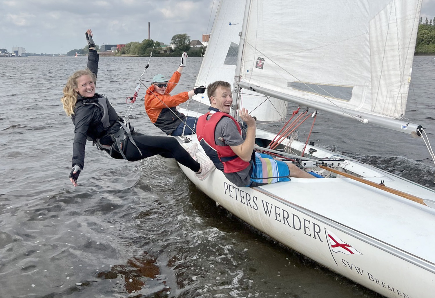 Drei Personen segeln lachend auf einer Jolle des Segelvereins Weser in Bremen. Eine Frau hängt im Trapez seitlich über dem Wasser, während die beiden anderen Crewmitglieder im Boot sitzen und in die Kamera winken. Das Boot trägt den Namen ‚Peters Werder‘ und fährt bei lebhaftem Wind über die Weser.