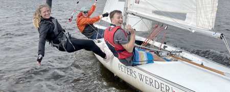 Drei Personen segeln lachend auf einer Jolle des Segelvereins Weser in Bremen. Eine Frau hängt im Trapez seitlich über dem Wasser, während die beiden anderen Crewmitglieder im Boot sitzen und in die Kamera winken. Das Boot trägt den Namen ‚Peters Werder‘ und fährt bei lebhaftem Wind über die Weser.